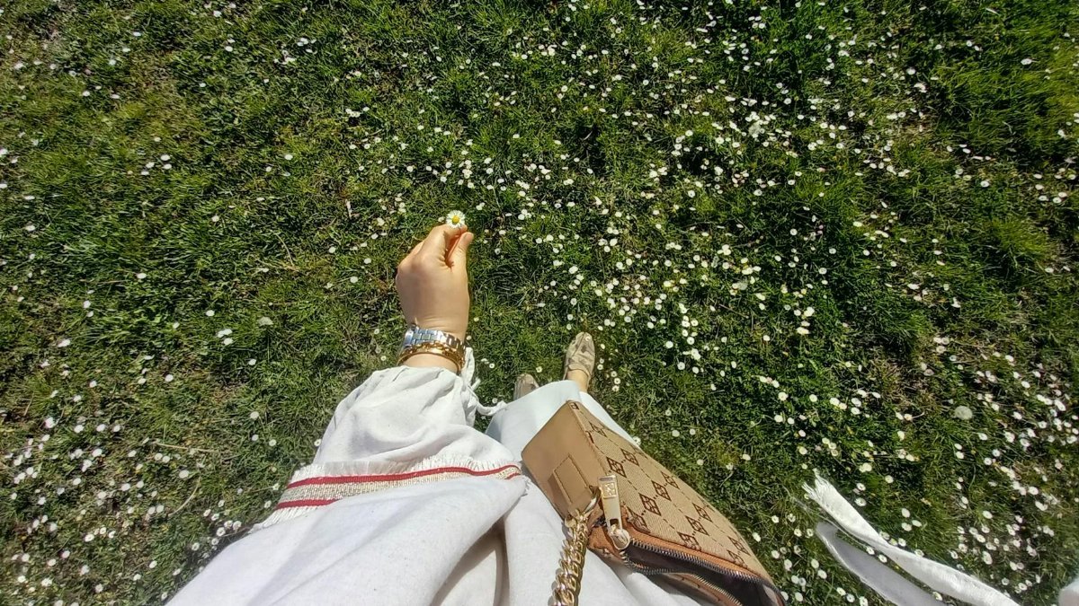 View from above of a woman in a white garment holding a daisy in a sunny, flower-covered meadow.