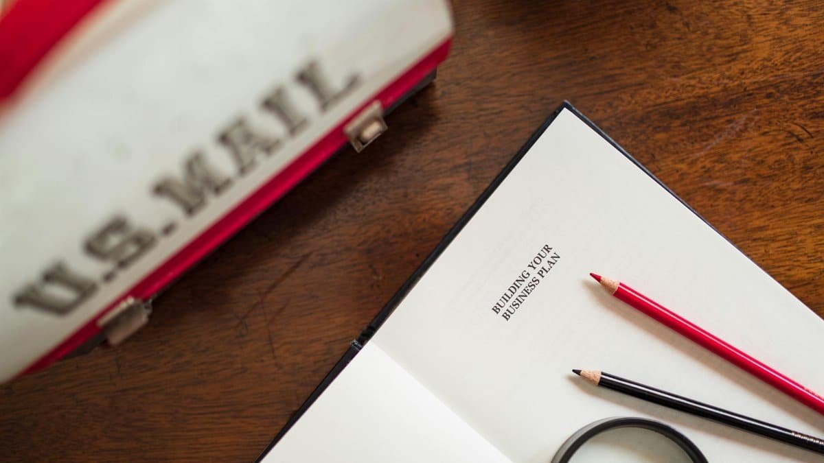 Open book with 'Building Your Business Plan' text, pencils, and U.S. Mail bag on a wooden desk.
