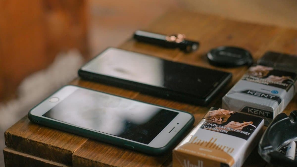 Close-up of mobile phones, cigarette packs, and gadgets on a wooden table.