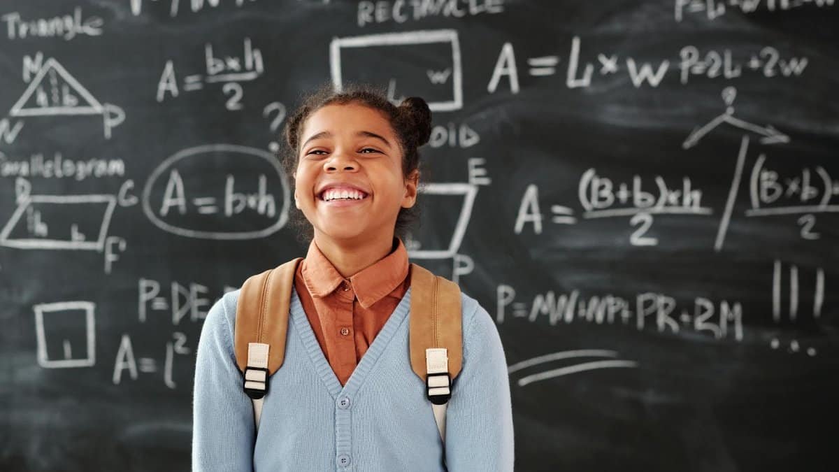 Smiling girl with backpack in a classroom, standing in front of a chalkboard with math formulas.