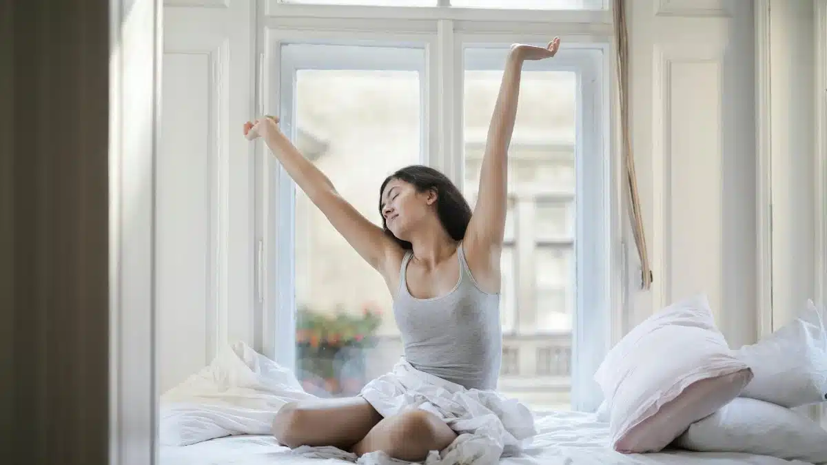 A woman in a gray tank top stretches joyfully on her bed in a bright room, embracing a new day.