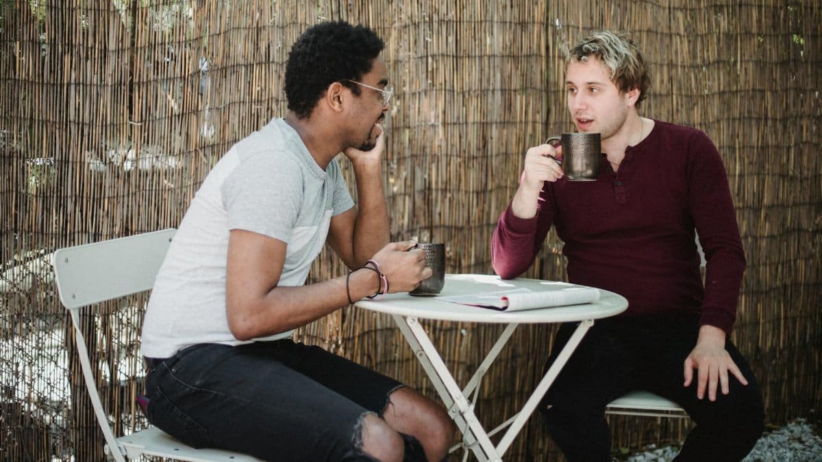 Two men enjoying a casual conversation over coffee outdoors with a rustic background.