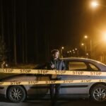 Man stands behind a car and stop tape at night, creating a mysterious scene.