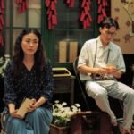 Two people in a vintage Asian room, surrounded by cultural decor, including a map and red decorations.