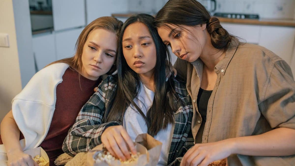 Three young women consoling each other while sharing popcorn indoors, signifying friendship and support.