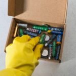 Yellow-gloved hand handling used batteries for safe recycling.