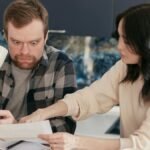 A couple reviews household bills in a kitchen, appearing concerned and focused.
