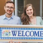 A cheerful couple stands at their new home entrance holding a welcome sign.