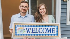 A cheerful couple stands at their new home entrance holding a welcome sign.