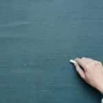 Hand holding chalk ready to write on a blank blackboard surface.