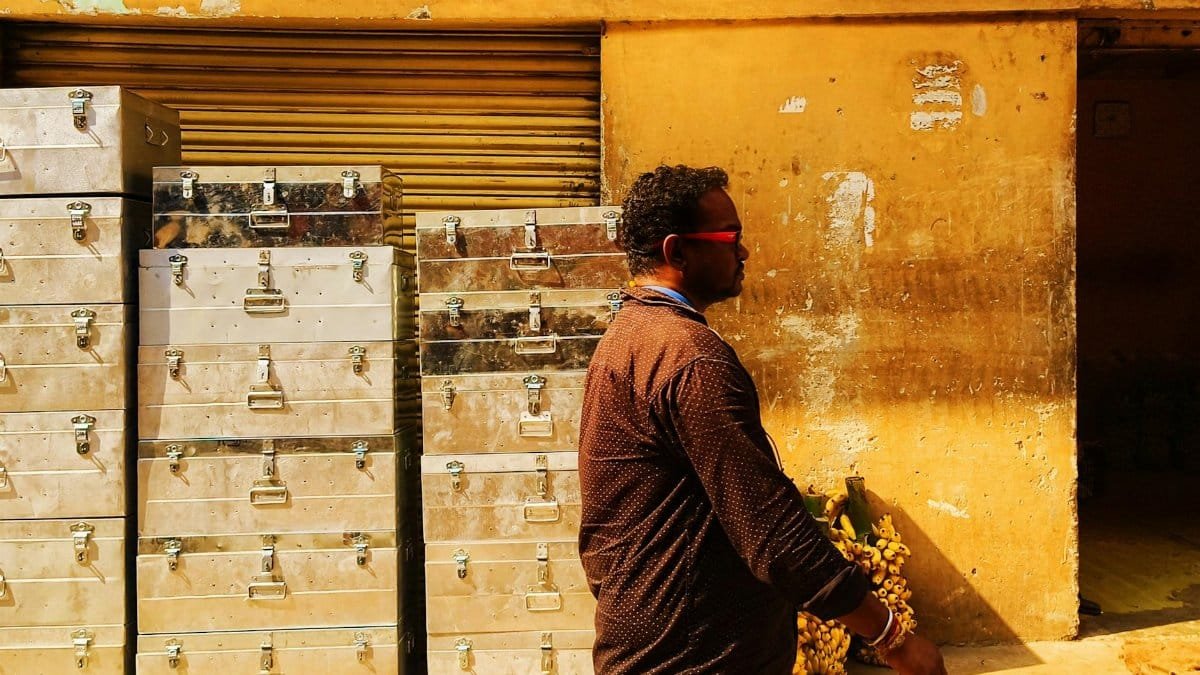 A man in sunglasses walks by metallic boxes outside a building in bright sunlight.