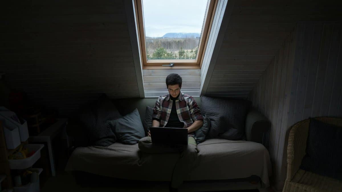 A young man focuses on his laptop in a dimly lit attic room with a window view.