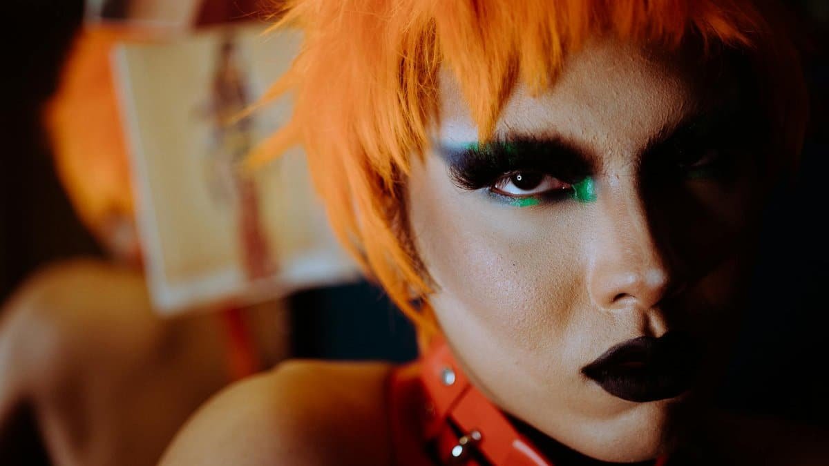 Expressive portrait of a young adult with vibrant orange hair and bold makeup, showcasing unique style and personality.