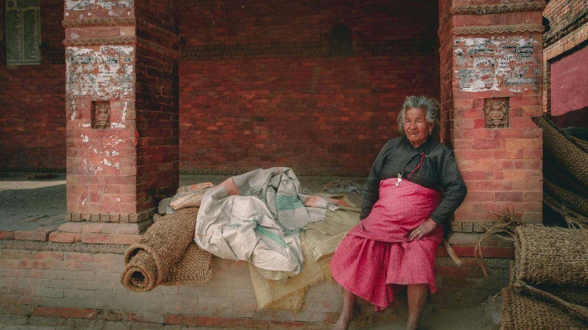 Elderly woman sitting in a traditional setting, weathered bricks and textiles.
