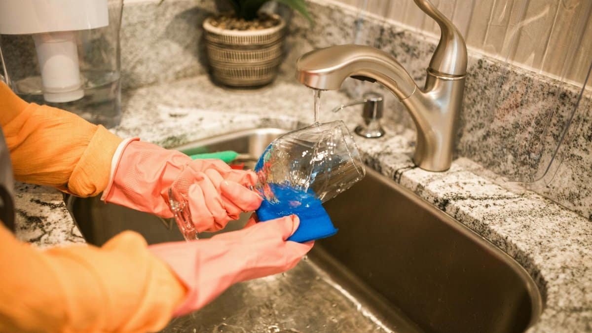 Close-up of hands wearing gloves washing a glass in a kitchen sink under a running faucet.