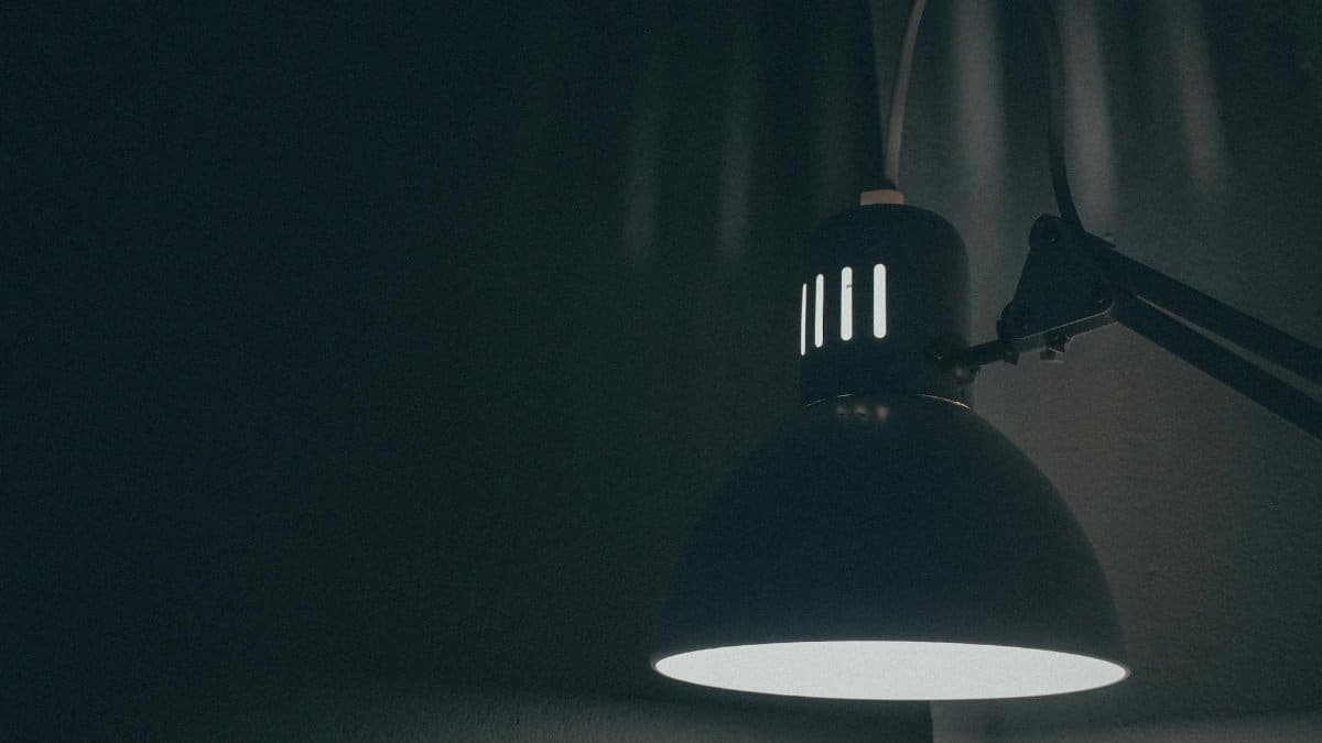 A dark, moody close-up of a lit desk lamp, showcasing warm ambient lighting in a minimalist setting.