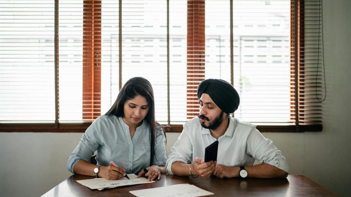 Indian woman with papers and pen writing on paper poster with ethnic Indian man in turban sitting nearby at wooden table
