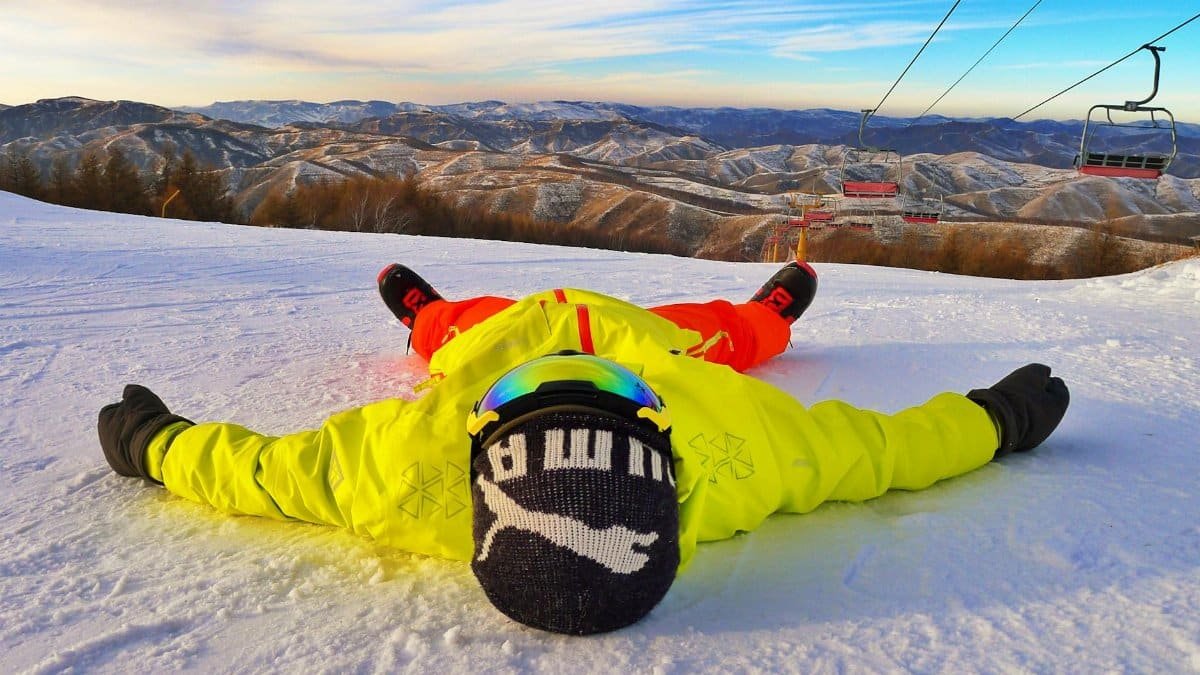 Person in vibrant winter clothing relaxing on a snowy mountain at a ski resort with a clear blue sky.