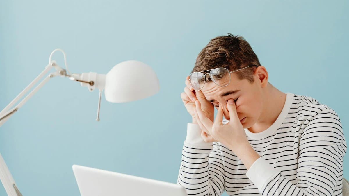 A young man rubs his eyes while working on a laptop, looking tired under a bright desk lamp.