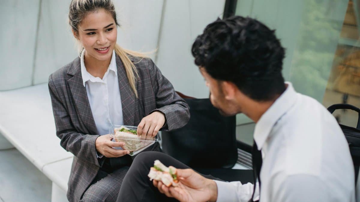 Two business professionals in formal attire enjoy a lunch break outdoors, sharing sandwiches and conversation.