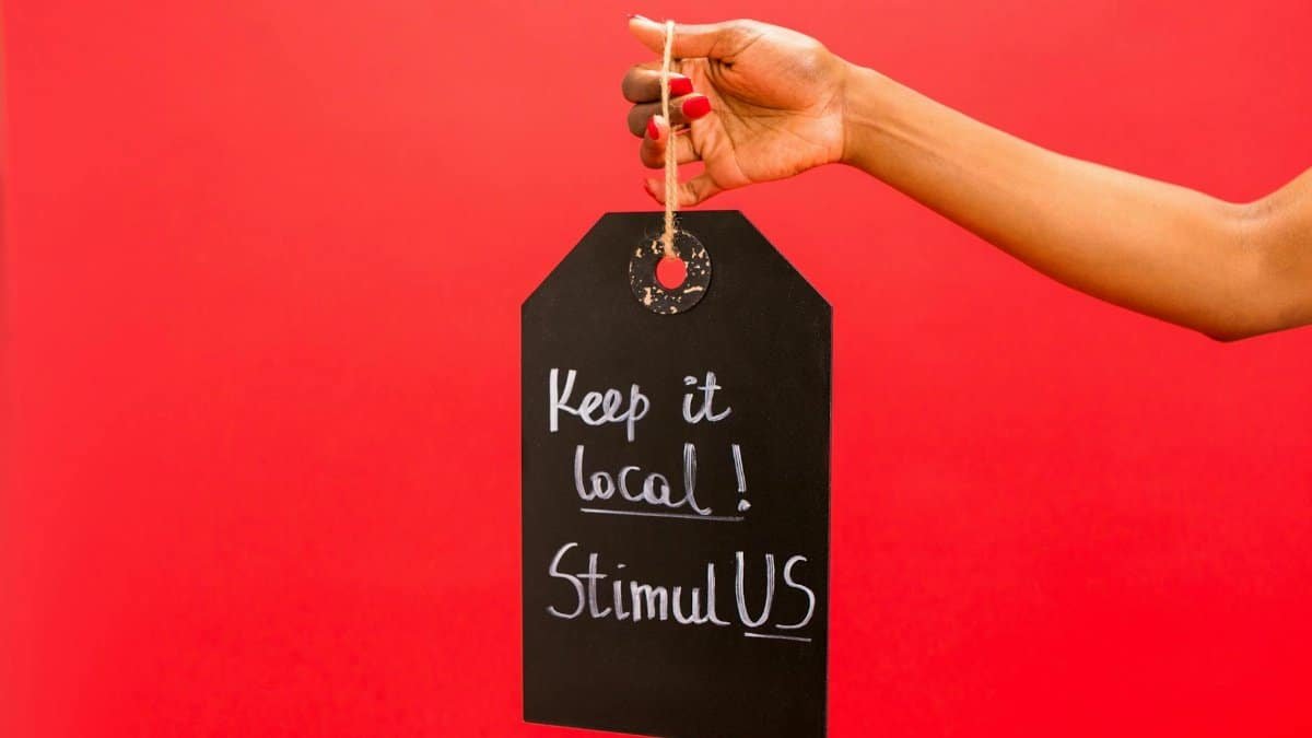 A hand presenting a chalkboard tag reading 'Keep it local! StimulUS' against a vibrant red backdrop.