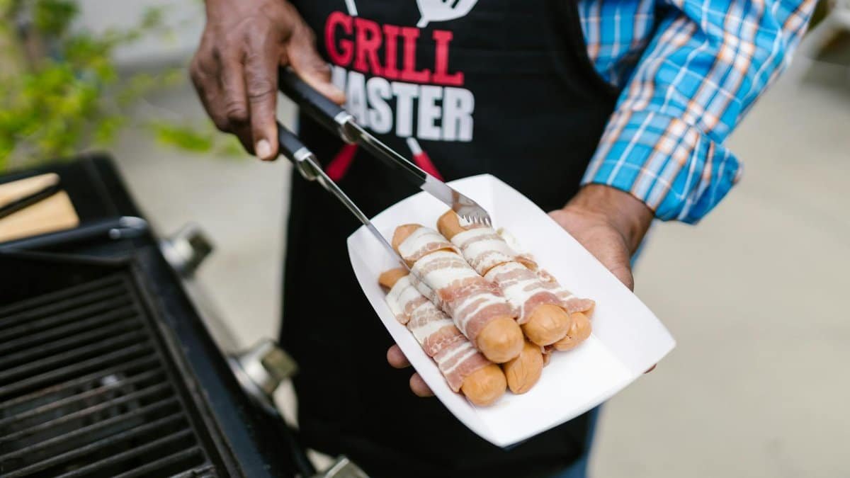 A person in an apron grilling bacon-wrapped hotdogs outdoors on a sunny day.