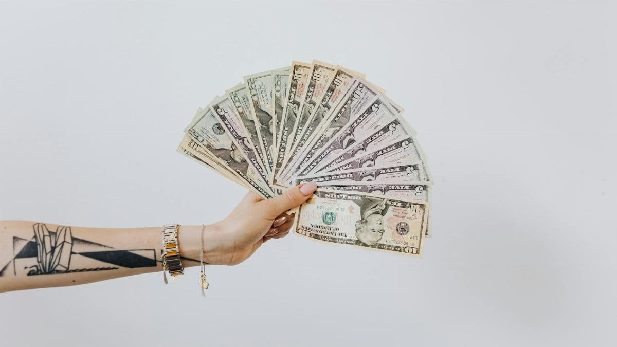 Tattooed arm holding a fan of US dollar bills on a white background, symbolizing wealth and financial success.