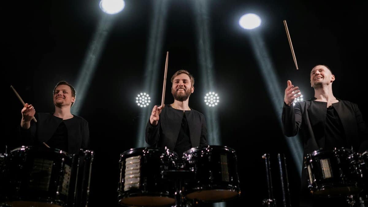 Three male drummers performing with drumsticks under vibrant stage lights.