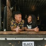 Two joyful business owners lean on a window sill with a 'Black Businesses Matter' sign visible.