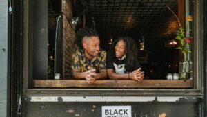 Two joyful business owners lean on a window sill with a 'Black Businesses Matter' sign visible.