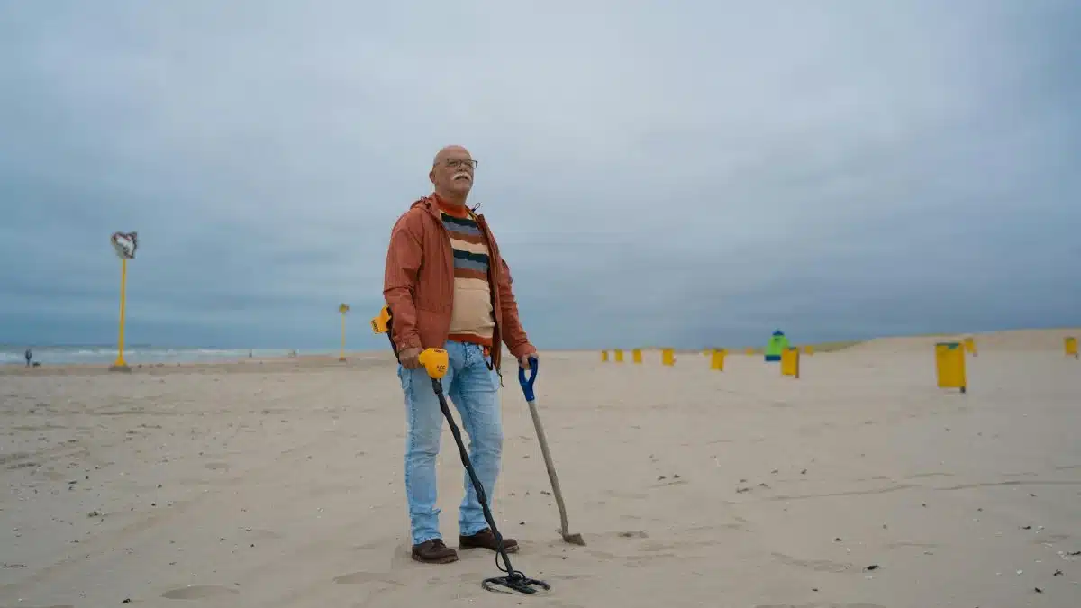 Elderly man with metal detector and shovel searching for treasures on a cloudy beach.
