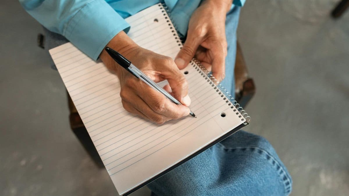 Close-up of hands writing in a spiral notebook with a pen, seated comfortably.