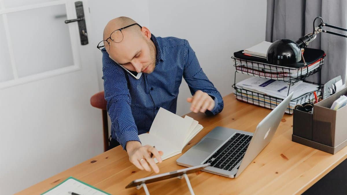 A man multitasking with phone, laptop, and notebook on his desk at home office.