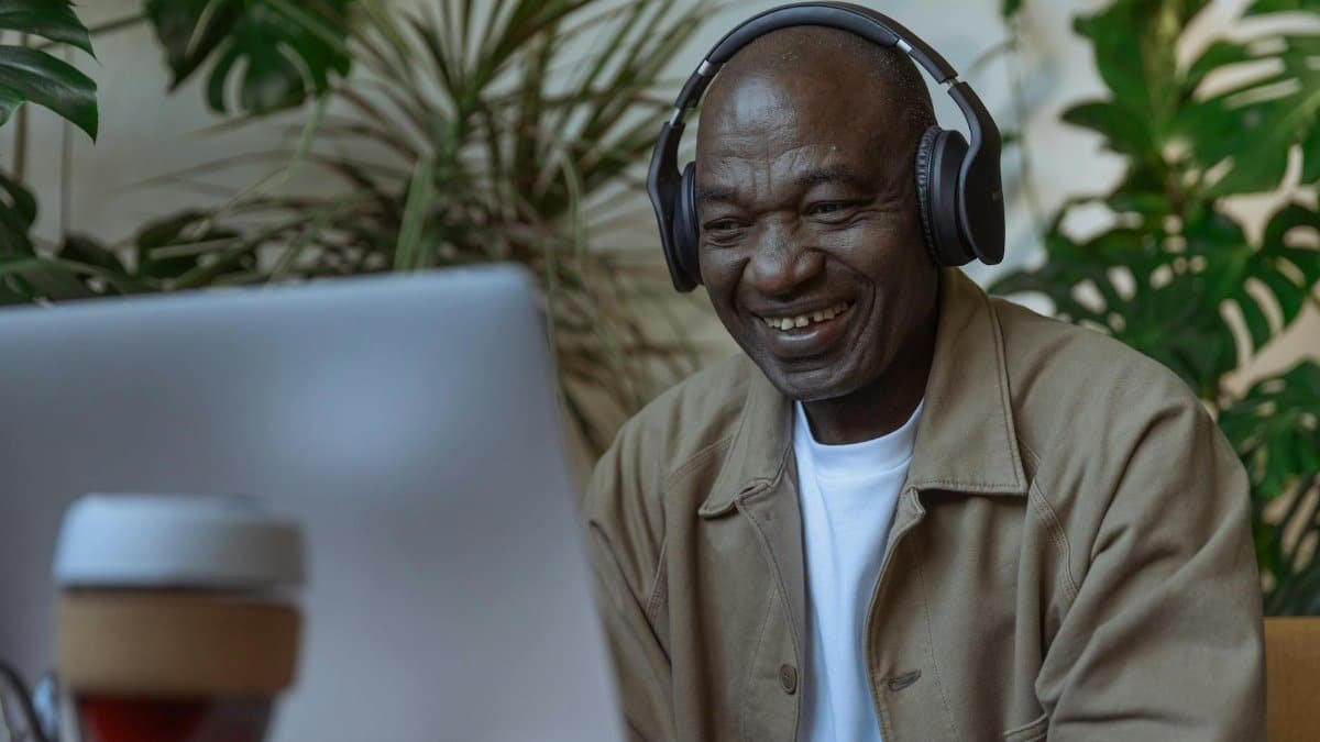 Senior man enjoying remote work on laptop with headphones in indoor setting with plants.