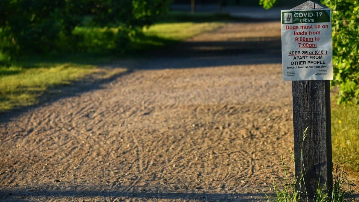 Signpost with COVID-19 safety rules on a park path, emphasizing social distancing and dog control.