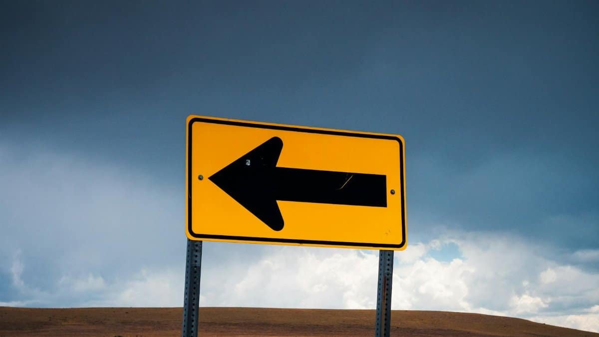 A bold yellow arrow sign against a dramatic overcast sky in a vast empty plateau landscape.