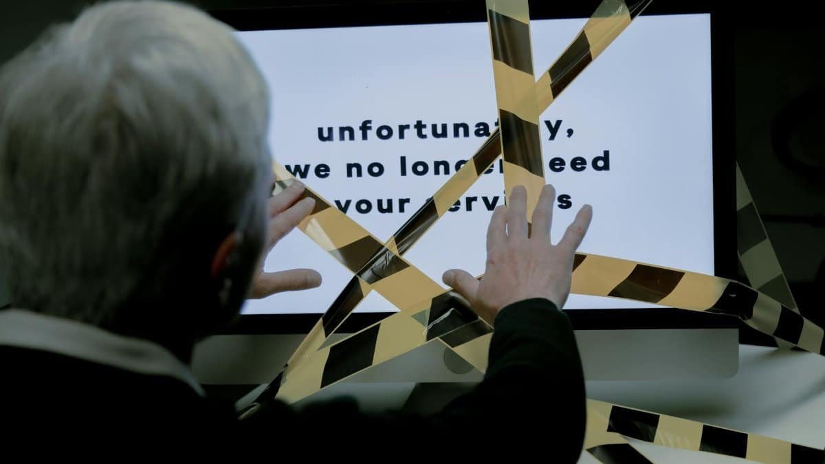 An elderly man reacts to a dismissal message on a computer, conveying ageism and unemployment themes.