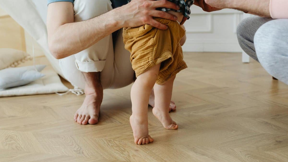 A heartwarming scene of a father helping his baby take first steps barefoot on a wooden floor.