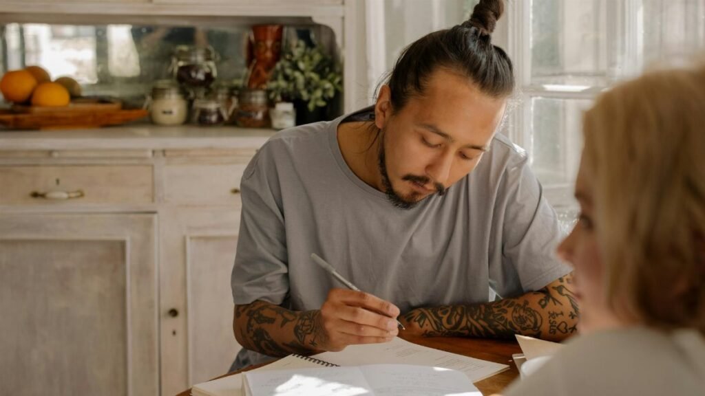 Tattooed young adults studying and taking notes at a table in a warmly lit room.