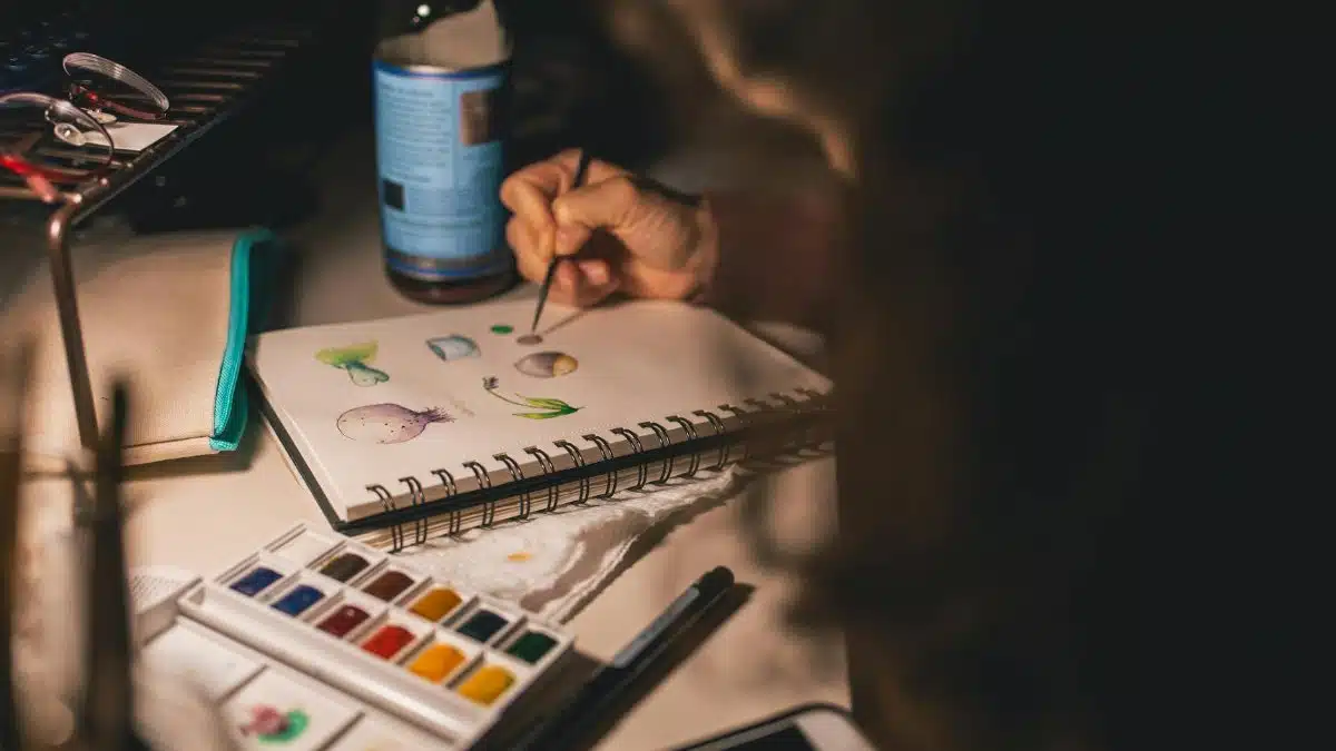 Artist painting with watercolors in a notepad at a desk. Creative process indoors.