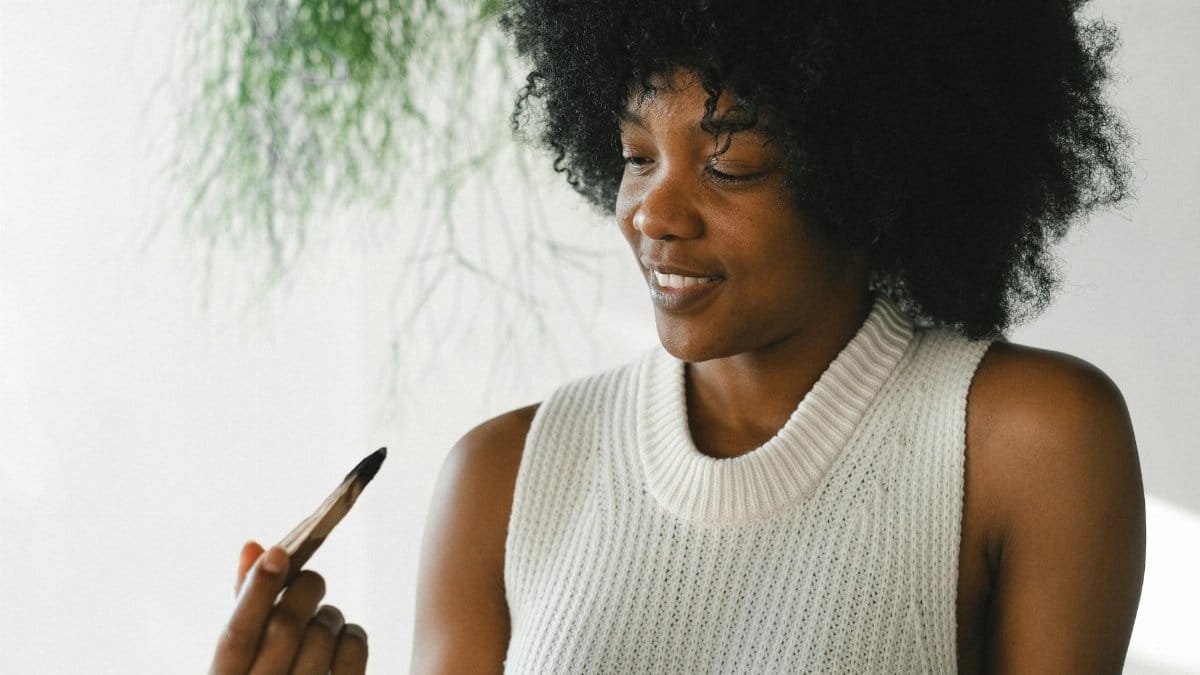 Satisfied African American female with wooden palo santo stick enjoying aromatherapy at home
