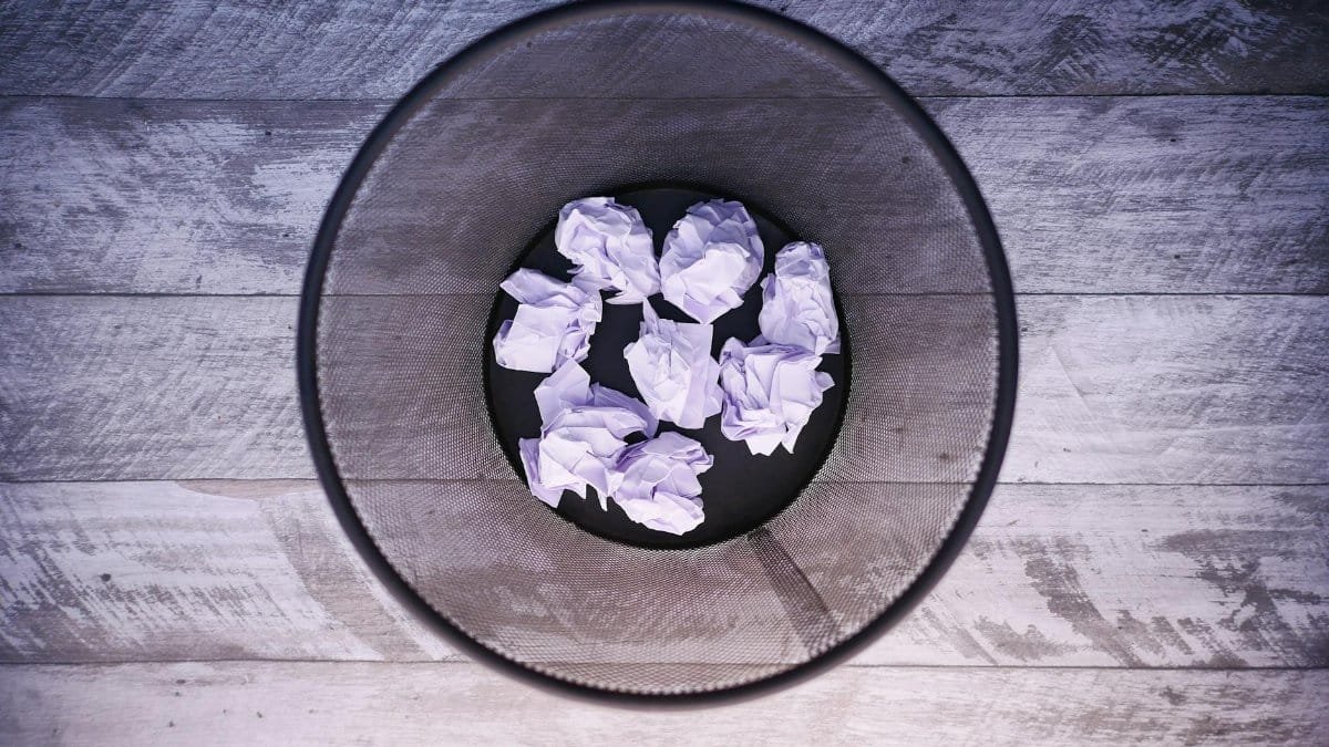 A top-down view of crumpled papers inside a black trash can on wooden flooring.