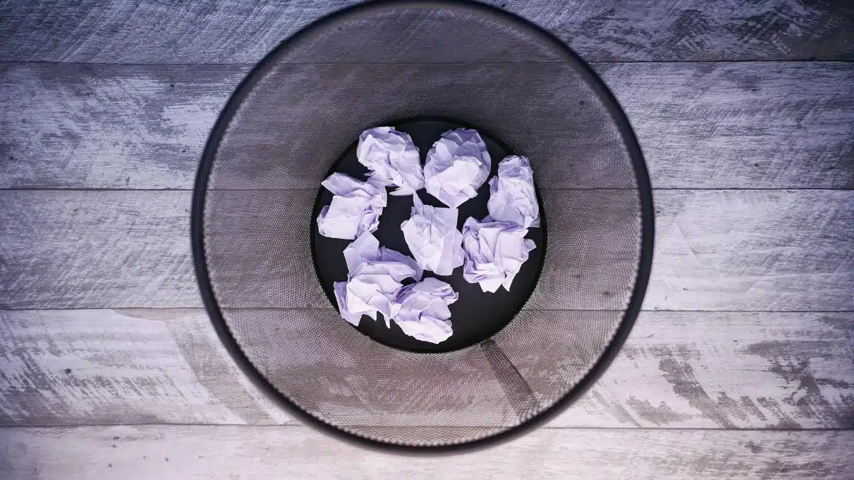 A top-down view of crumpled papers inside a black trash can on wooden flooring.