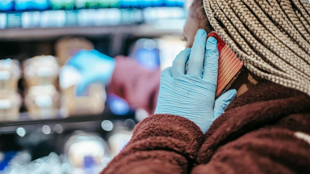 Side view of crop African American female customer in latex gloves with braids having phone call while picking food from shelf in grocery store