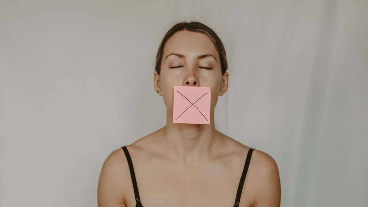 Young slender woman with closed eyes and mouth covered with sticky note showing cross on white background