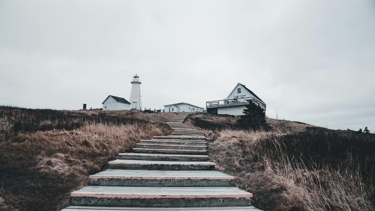 Moody view of a lighthouse atop a hill with steps leading up in a cloudy coastal setting.