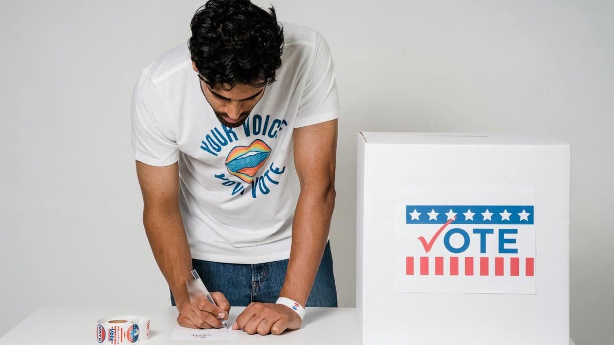 A man writes on a paper to cast his vote next to a ballot box in a studio setting.