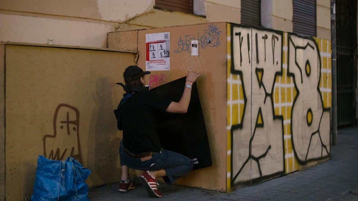 A street artist squats while adding graffiti to a wall on an urban street corner.