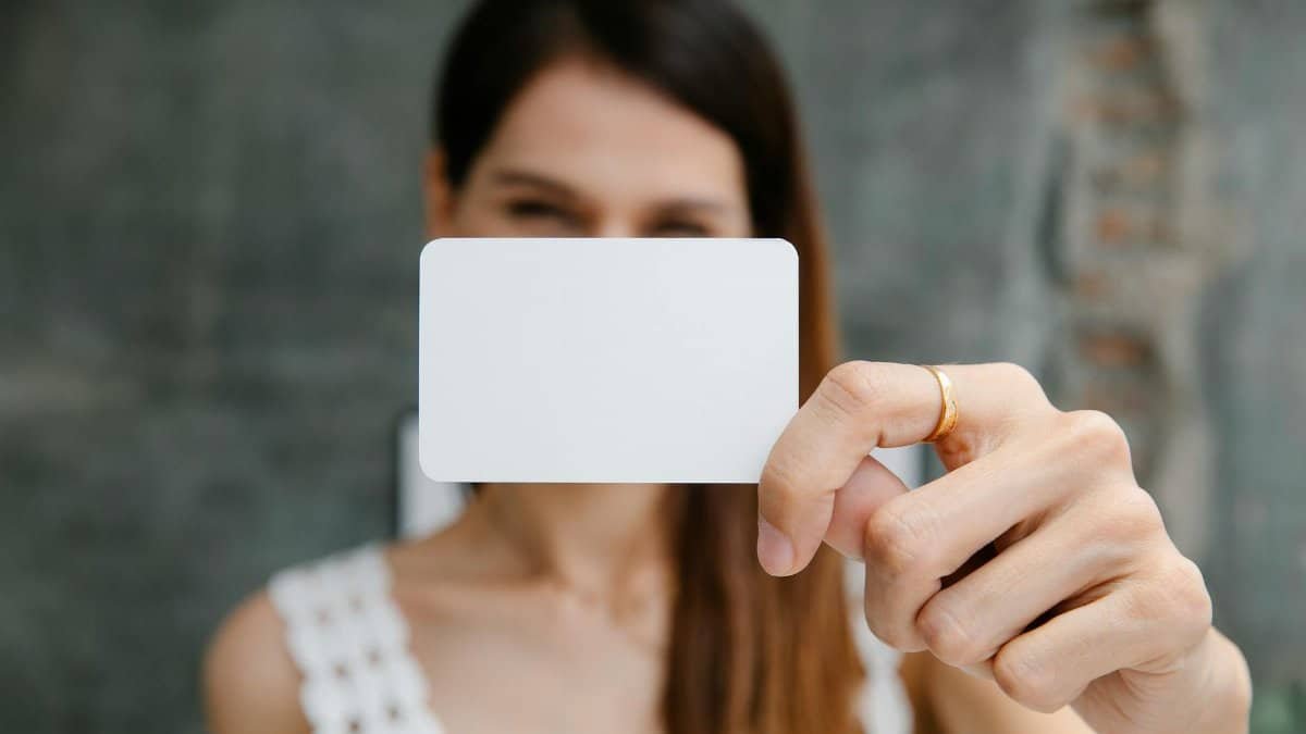 Young blurred female showing white blank business card and looking at camera in light room