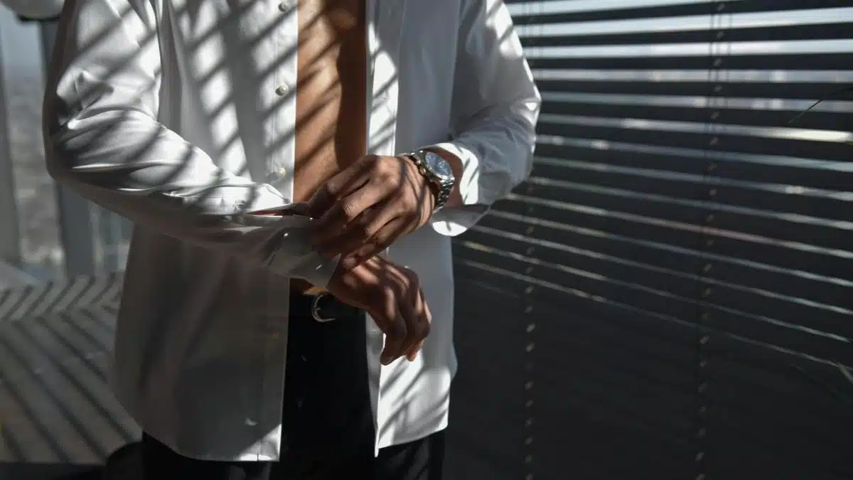 A fashionable man adjusting his shirt cuffs in sunlight filtered through blinds in a modern interior space.